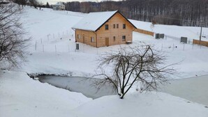 A wooden cabin with a sloped roof, surrounded by snow-covered ground and a small stream.