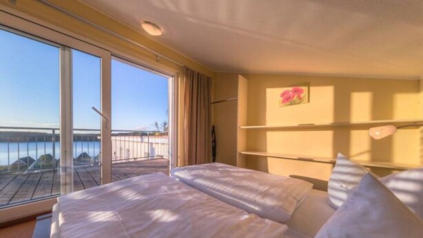 Habitación de hotel con un amplio ventanal que ofrece vista a un cuerpo de agua y un muelle. Hay dos camas con ropa blanca y un pequeño aparador contra la pared.