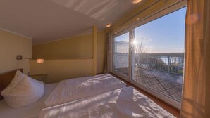 Habitación de hotel con un ventanal grande, una cama con ropa de cama blanca y una mesita de noche con lámpara.