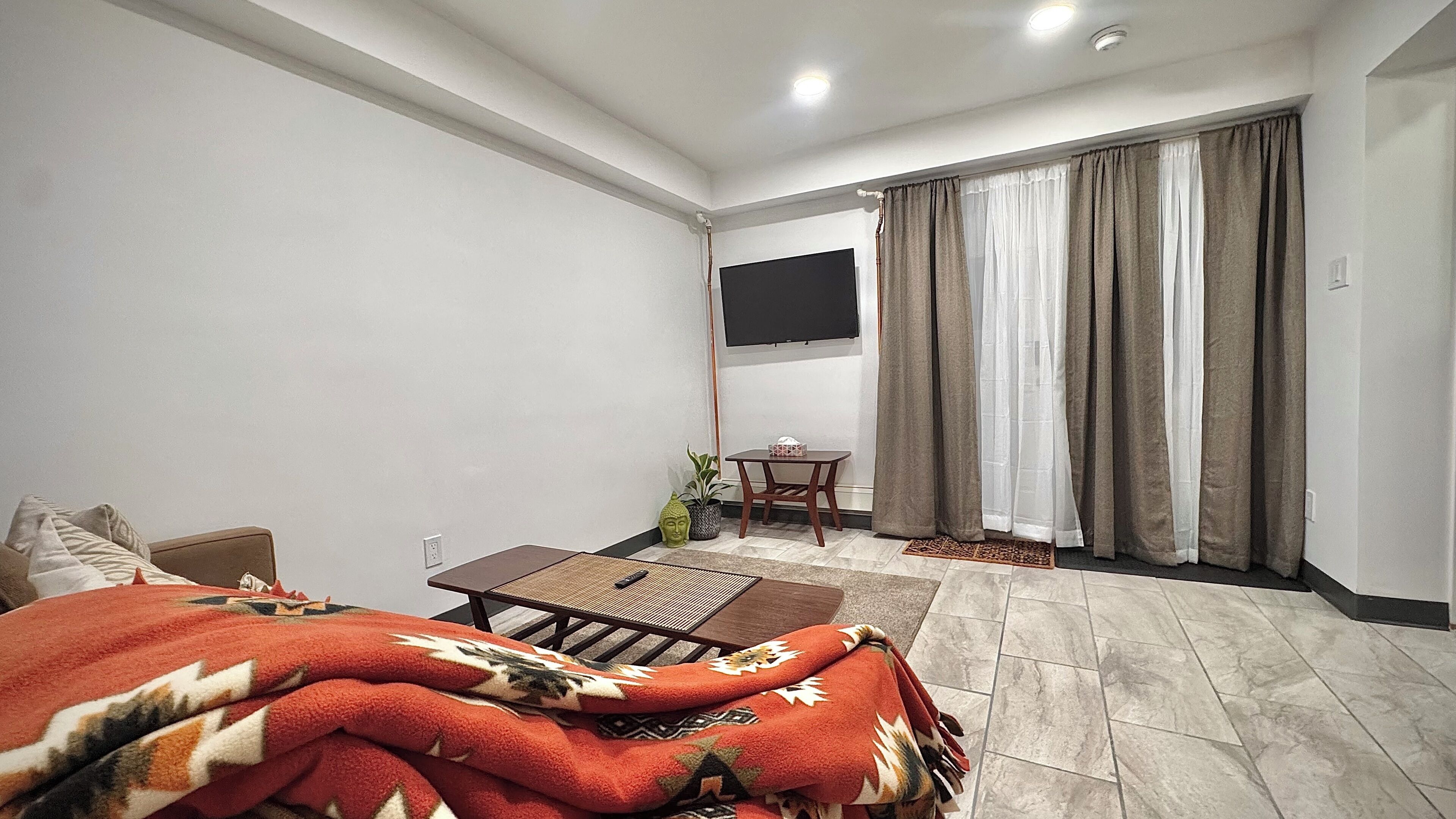 A modern hotel room with a bed, a small table, a chair, a TV mounted on the wall, and a sliding door leading to a balcony.
