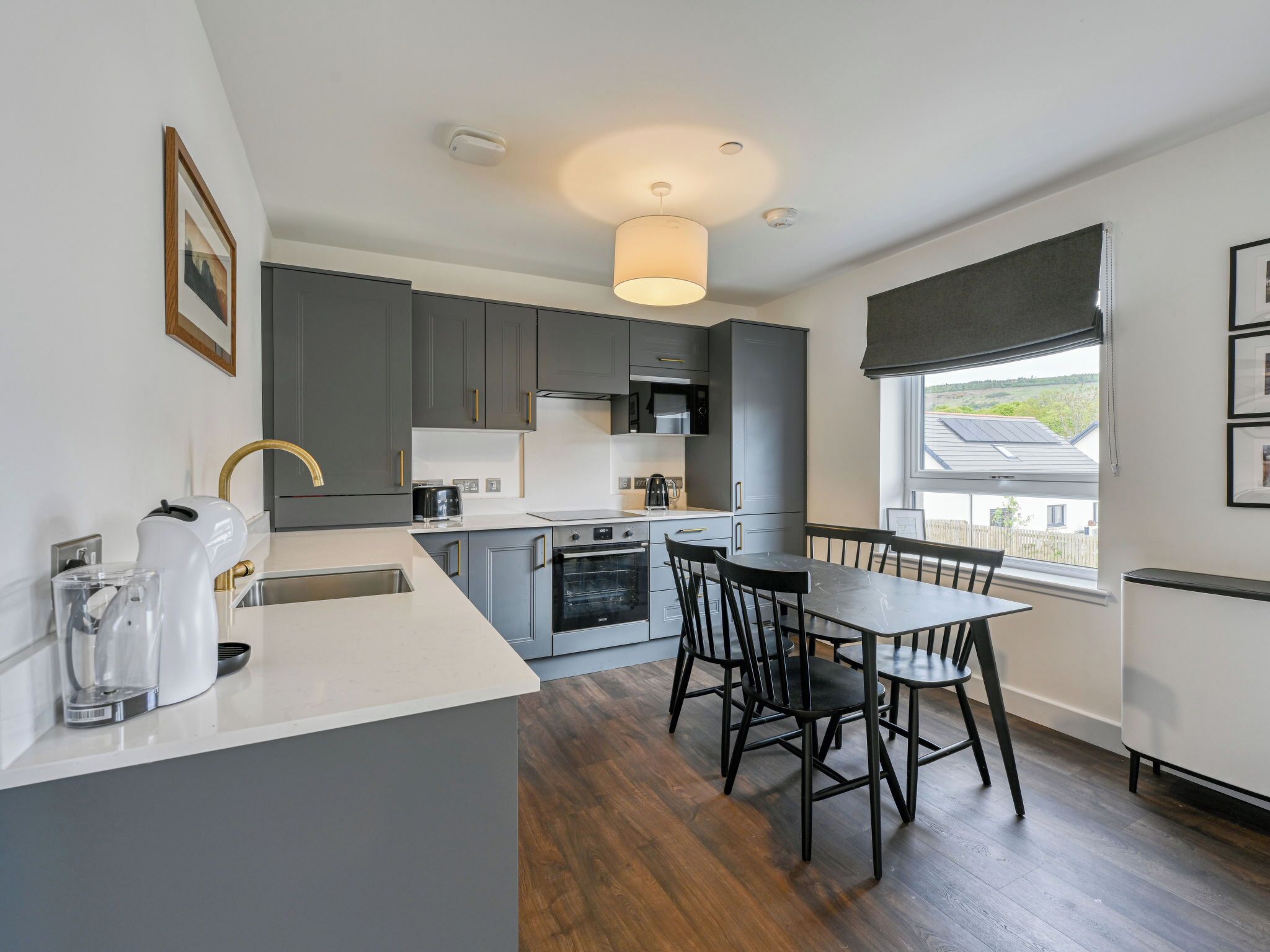 A modern kitchen with grey cabinets, a dining table with chairs, a coffee maker, and a view of the outside through a window.