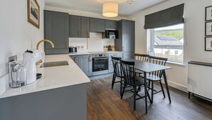 Uma cozinha moderna com armários cinzentos, uma mesa de jantar com cadeiras, uma cafeteira e vista para o exterior através de uma janela.