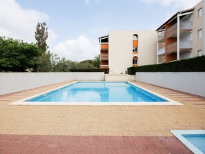 A rectangular pool with a tiled border, surrounded by a brick walkway. In the background, there is a multi-story building with balconies and a flat roof.