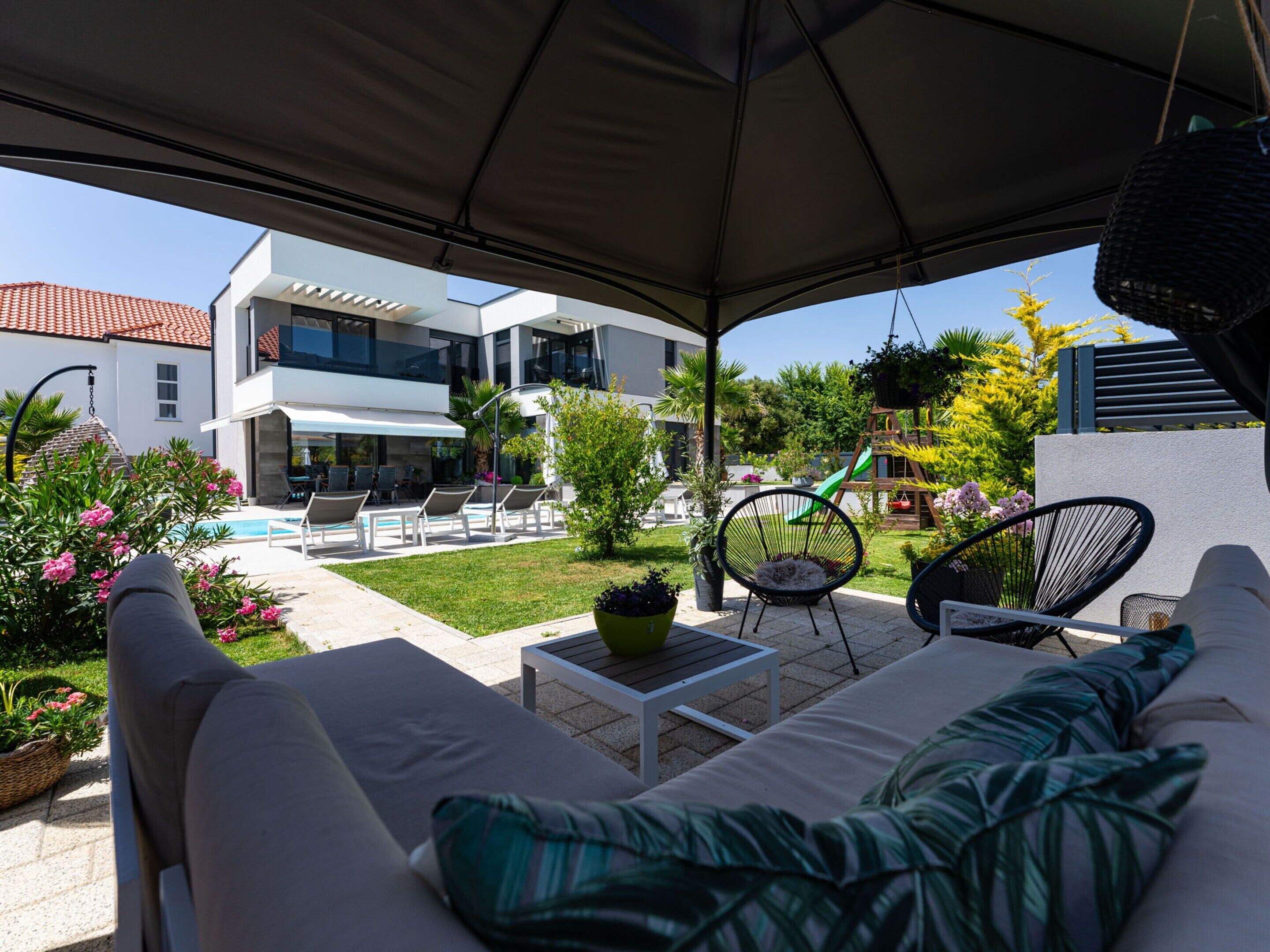 A covered outdoor seating area with a pool, lounge chairs, and a modern building in the background.