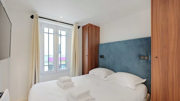 A hotel room with a bed, a wooden wardrobe, a blue accent wall, and a window with curtains.