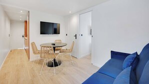 A modern, well-lit room with a round dining table, wicker chairs, a blue sofa, and a flat-screen TV mounted on the wall.