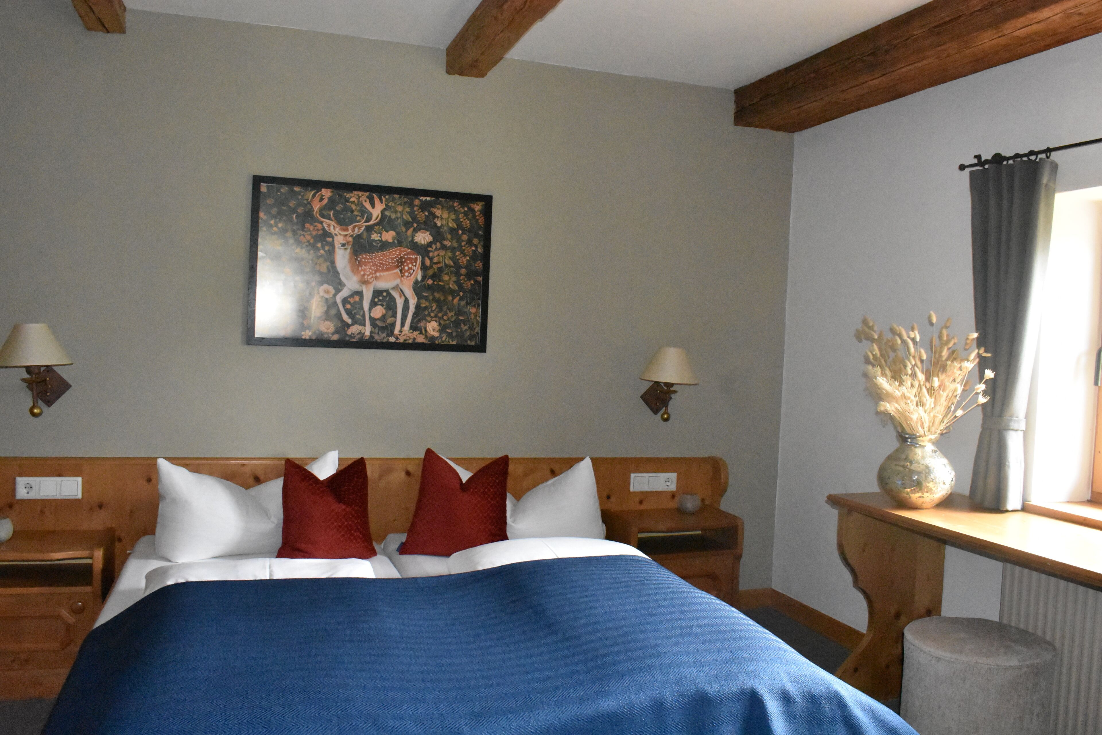 A bedroom with a bed, wooden headboard, two bedside tables with lamps, a nightstand, a wall-mounted painting of a deer, and a window with a curtain.