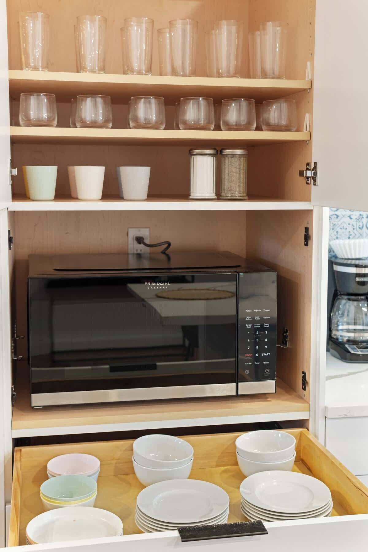 A kitchen with a microwave, shelves with glassware, and a drawer with plates.