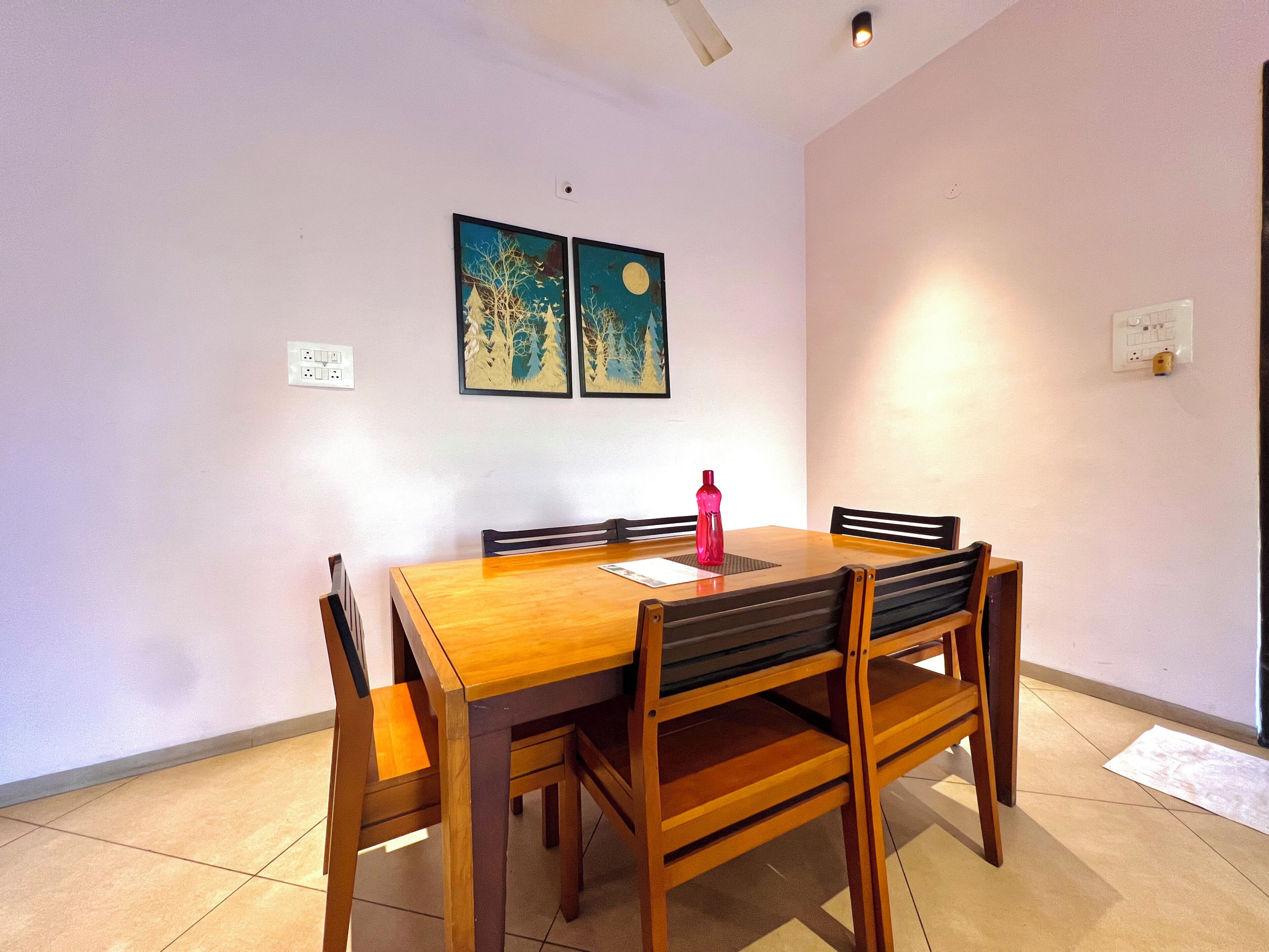A dining area with a wooden table and chairs, two wall-mounted paintings, and a red water bottle on the table.