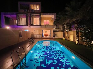 A modern two-story house with a swimming pool, outdoor seating, and a well-lit entrance.