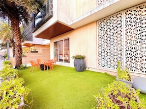 A modern building with a glass door, a decorative white lattice, and a small patio with artificial grass and orange chairs.