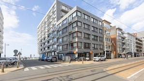 A modern multi-story building with a ground-floor commercial space, a tram line, and a pedestrian crossing. - Spacious Studio Near Seafront (Middelkerke)