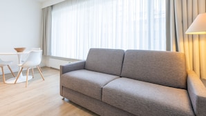 A modern living room with a grey sofa, a small round table with chairs, and sheer curtains on the window. - Spacious Studio Near Seafront (Middelkerke)