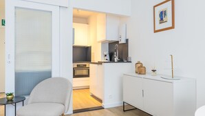 A modern kitchen with white cabinetry, a built-in oven, and a small dining area with a chair and table. - Spacious Studio Near Seafront (Middelkerke)