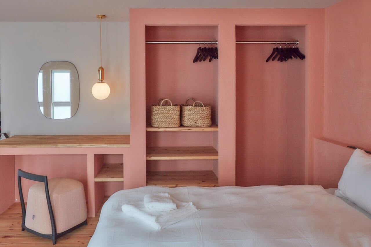 Un dormitorio con un escritorio de madera, un espejo, una lámpara colgante, un armario con ropa colgada y una cama con ropa de cama blanca.