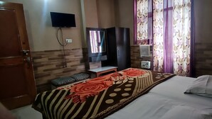 A hotel room with a bed, a TV mounted on the wall, a wooden wardrobe, and a window with floral curtains. - Hotel Ishaan International (Reasi)