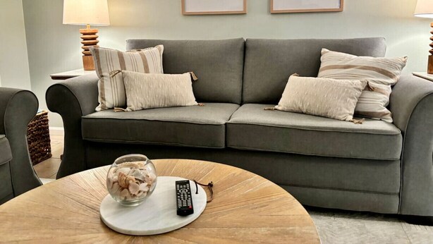 A living room with a grey sofa, two framed pictures on the wall, a round wooden coffee table with a remote control and decorative item, and two lamps on either side of the sofa. - Cozy Beach Condo! Safe Complex! (Panama City Beach)
