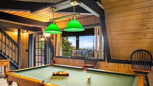 Une table de billard recouverte d’un drap vert, deux chaises et une fenêtre donnant sur des arbres et des montagnes.