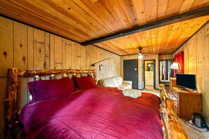 Ein Holzblockhaus-Interieur mit einem Bett, einem Sessel, einem Fernseher und einem durch eine offene Tür einsehbaren Badezimmer.