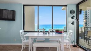 A room with a large window overlooking the ocean, a white table with chairs, and a small balcony with a railing. - Ocean Front Beach Condo (Panama City Beach)