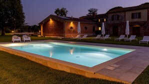 A rectangular swimming pool with a tiled edge, surrounded by a grassy area. There are white lounge chairs and umbrellas arranged around the pool. A brick building with a sloped roof and stone walls is visible in the background. - Tenuta Francigena 8 Emma Villas (Castelfranco di Sotto)