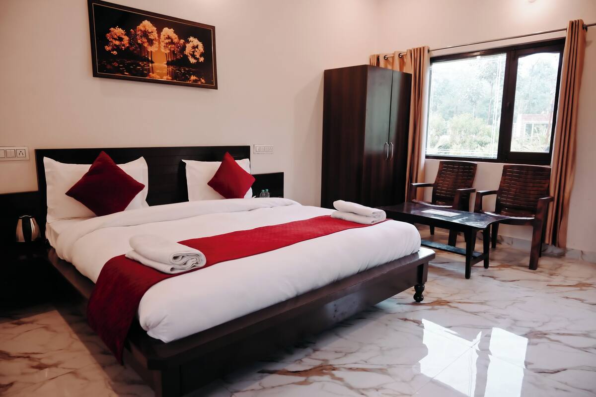 A neatly made bed with white and red linens, a wooden headboard, and a red throw blanket. There is a wooden wardrobe and a table with chairs in the room.