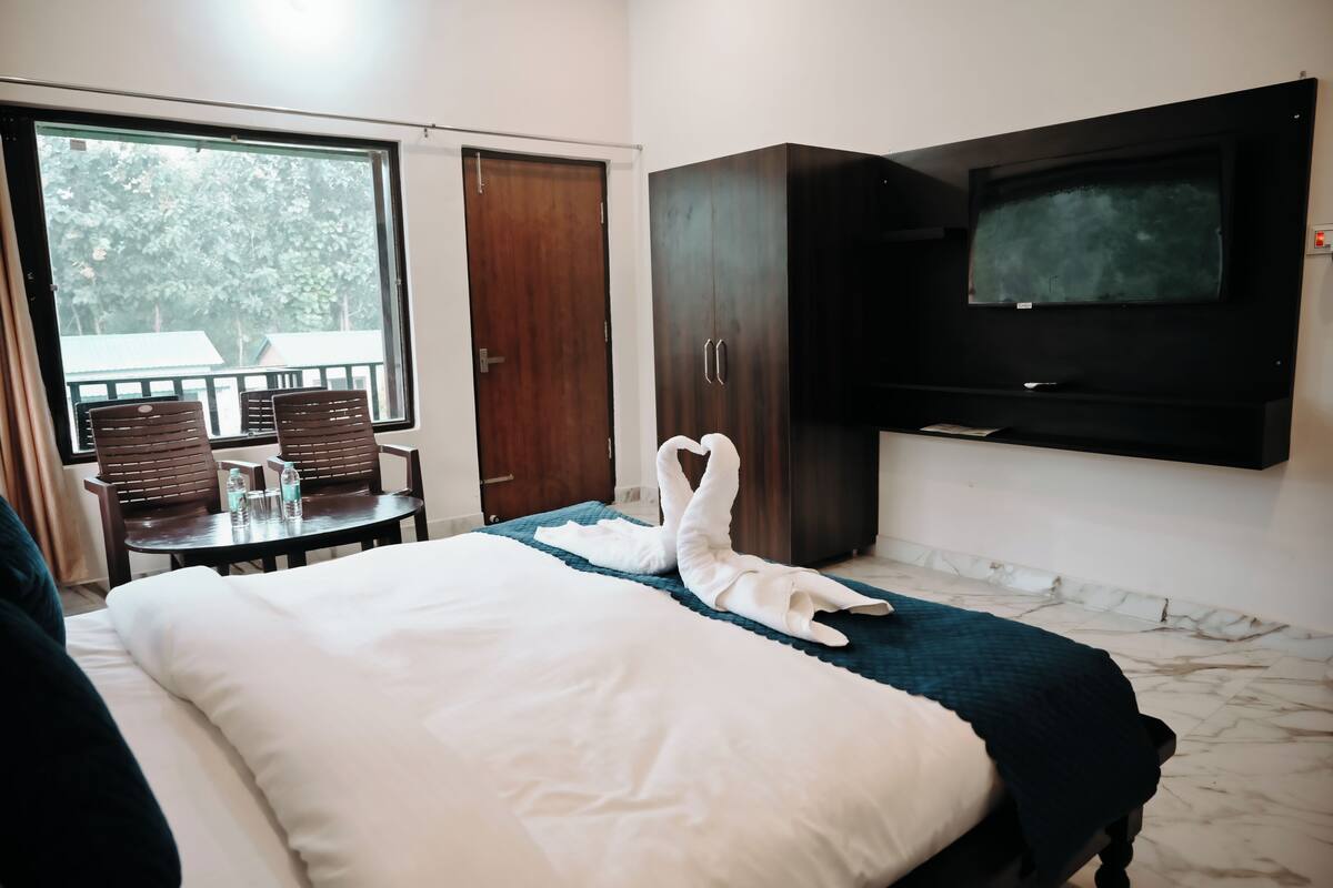 A hotel room with a bed, a TV, a wardrobe, a small table with chairs, and a window with a view of trees.