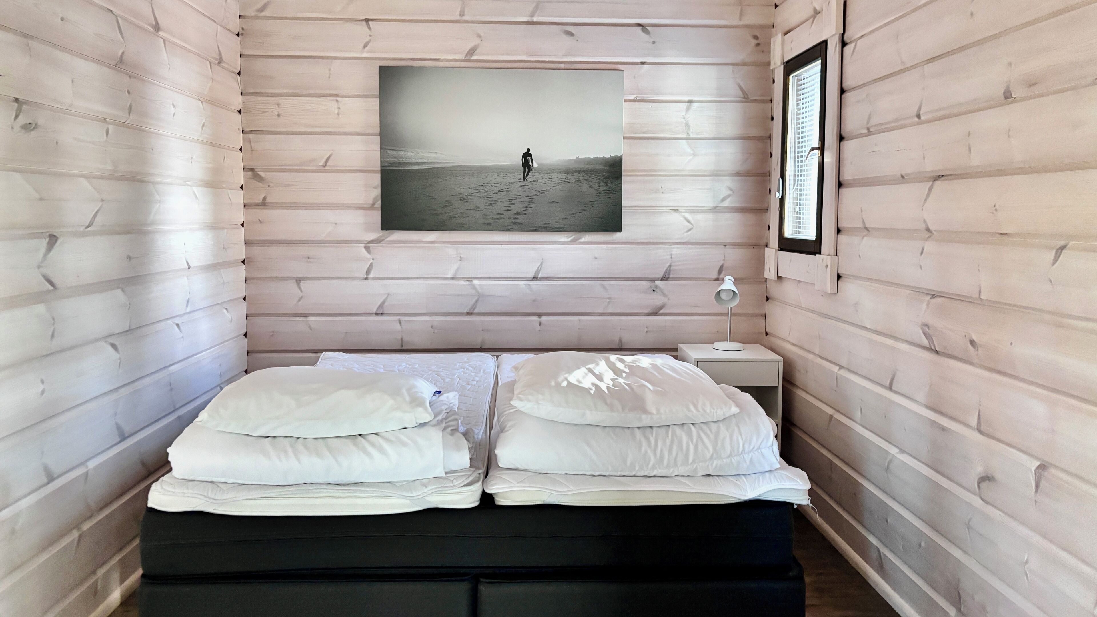 A cozy room with a bed, a nightstand, a framed picture of a beach, and a window with blinds.