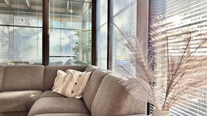 Un salon moderne avec un canapé d’angle beige, une plante décorative et de grandes fenêtres équipées de stores.