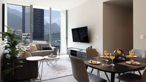 A modern hotel room with a large window, a sofa, a dining table set for four, and a flat-screen TV mounted on the wall.