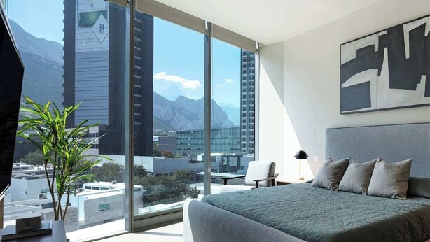 A modern bedroom with a large bed, a view of mountains, and a cityscape through floor-to-ceiling windows.