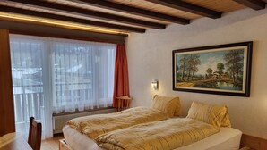 A bedroom with a wooden bed, a wooden nightstand, a chair, a window with sheer curtains, a radiator, and a painting on the wall. - Apartments Atlas (Saas Grund)