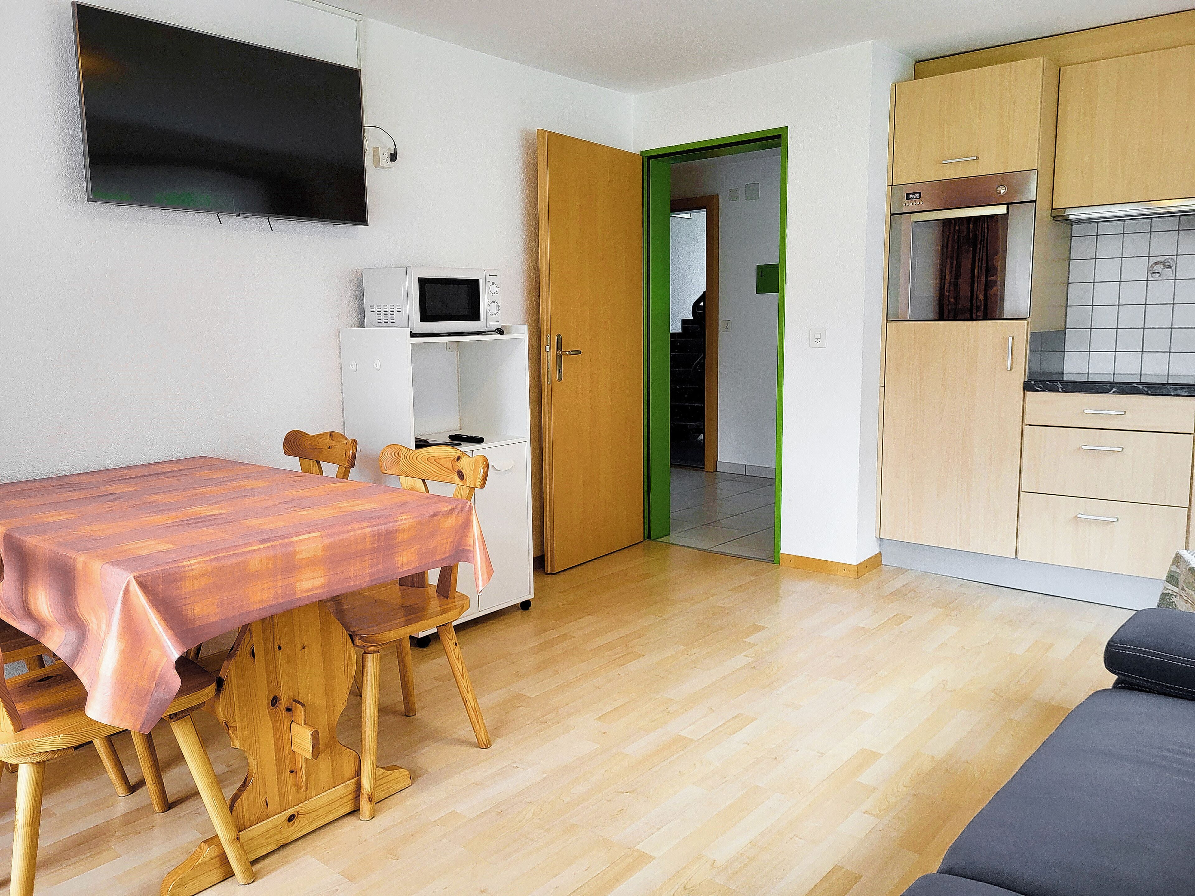 Una cocina compacta con comedor, armarios de madera, microondas y televisor montado en la pared.