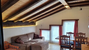 A cozy living room with a wooden ceiling, a sofa, a dining table with chairs, and a window with curtains. - Apartments Atlas (Saas Grund)