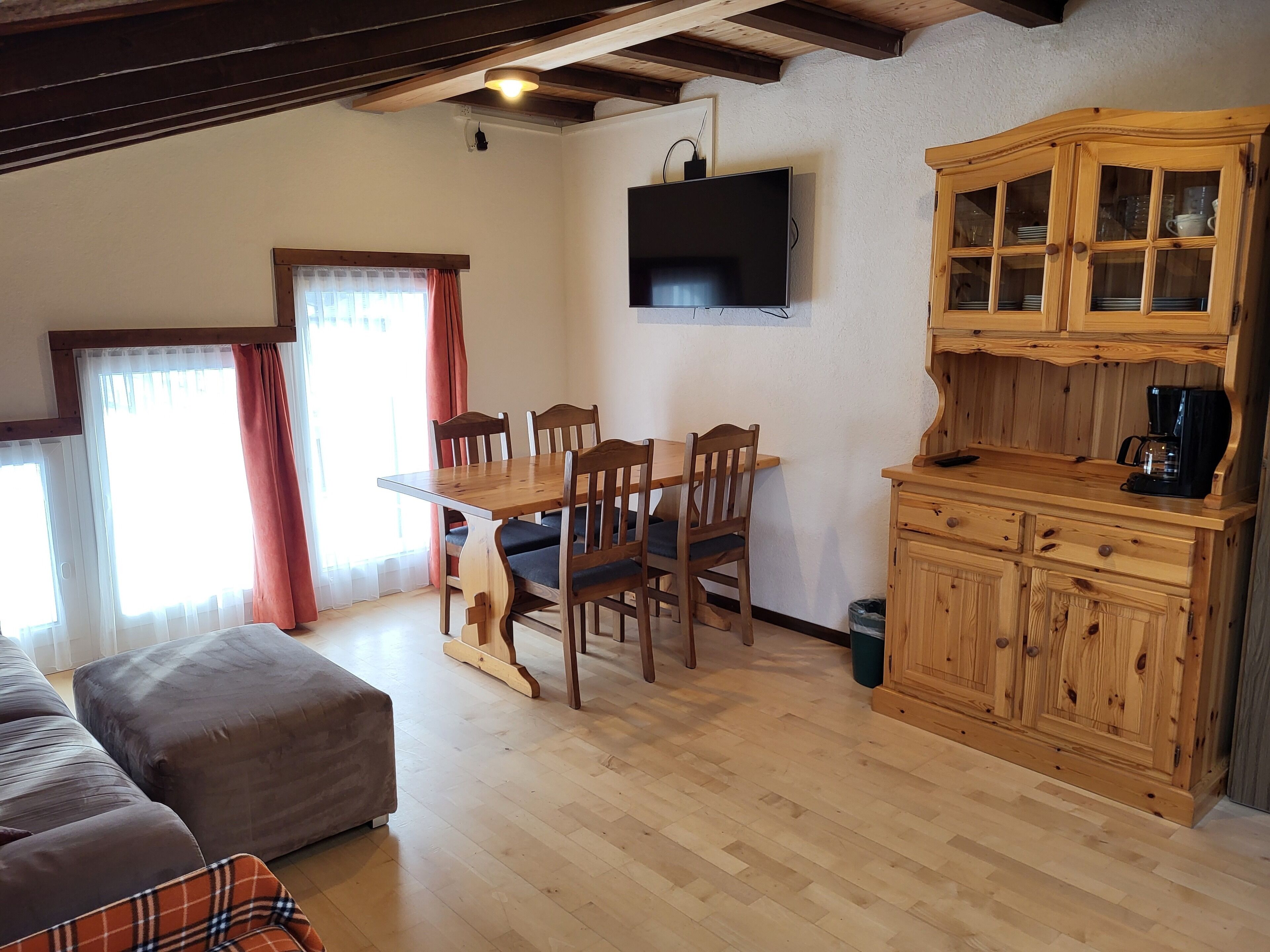 Una sala acogedora con una mesa de comedor de madera, sillas y un sofá. Un televisor está montado en la pared, y hay un mueble de madera con cajones.