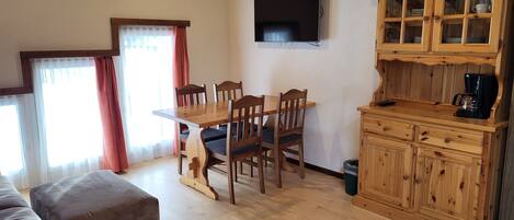 Una sala acogedora con una mesa de comedor de madera, sillas y un sofá. Un televisor está montado en la pared, y hay un mueble de madera con cajones.