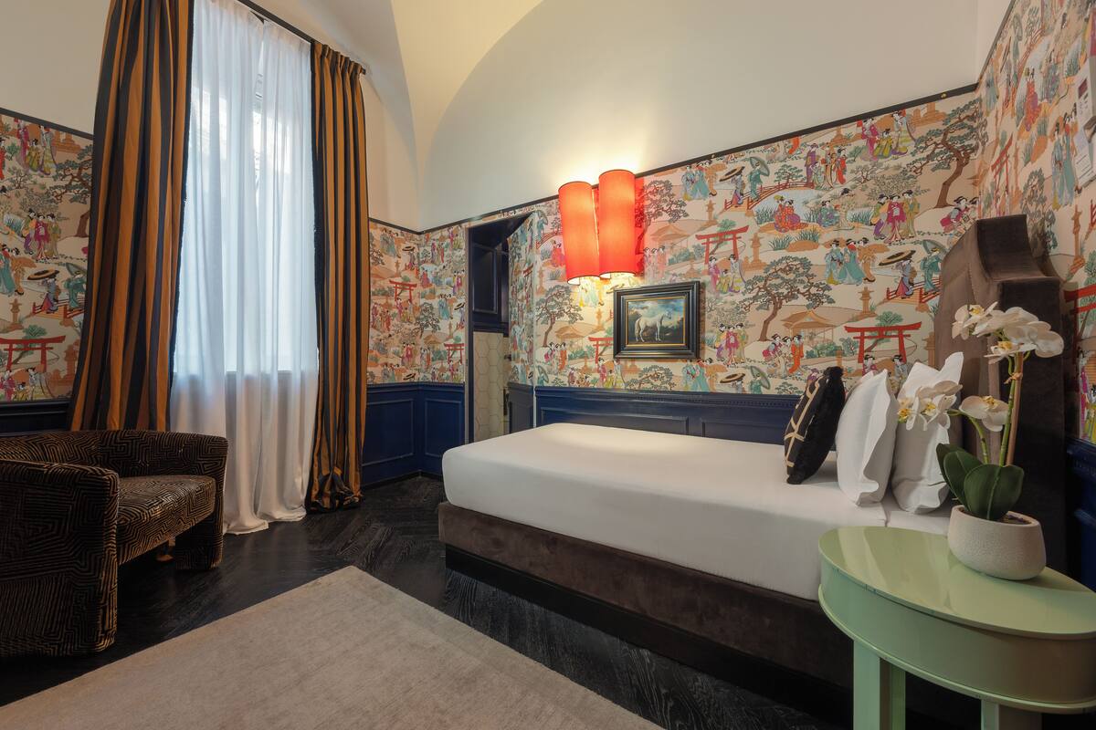 A hotel room with a bed, a chair, a small table, and a large window with curtains.