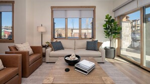 A modern living room with a beige sofa, two brown armchairs, a coffee table with a bowl and books, and large windows with a view of buildings and trees. - Downtown Fraser| Hot tub |Walkable (Fraser)
