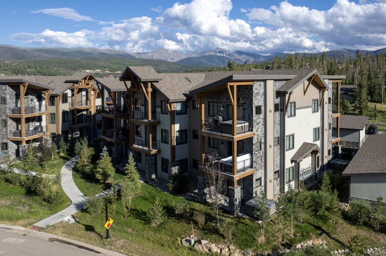 A modern multi-story residential building with stone and wood accents, balconies, and a view of mountains.