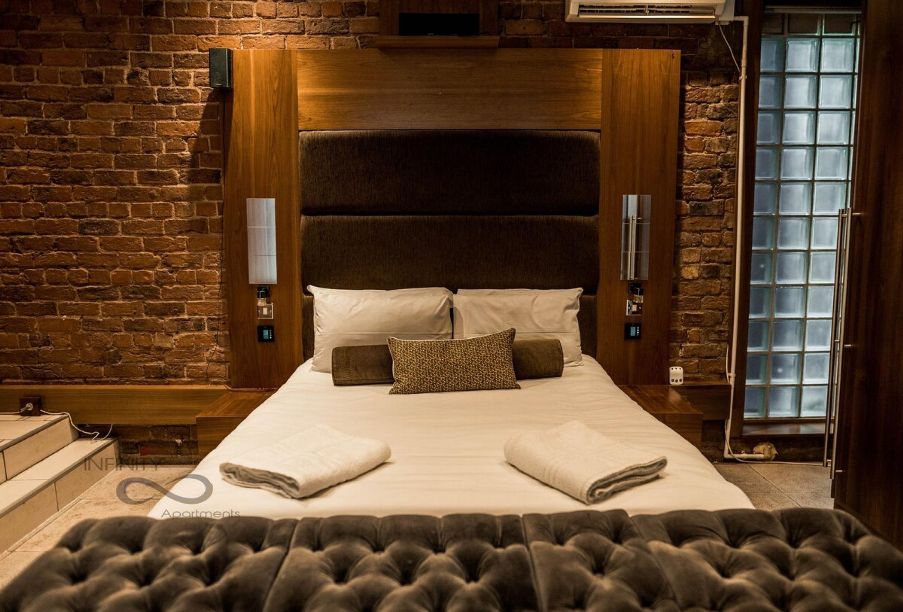 A hotel room with a brick wall, a wooden headboard, a large bed with pillows and a blanket, a small table, and a window with frosted glass.