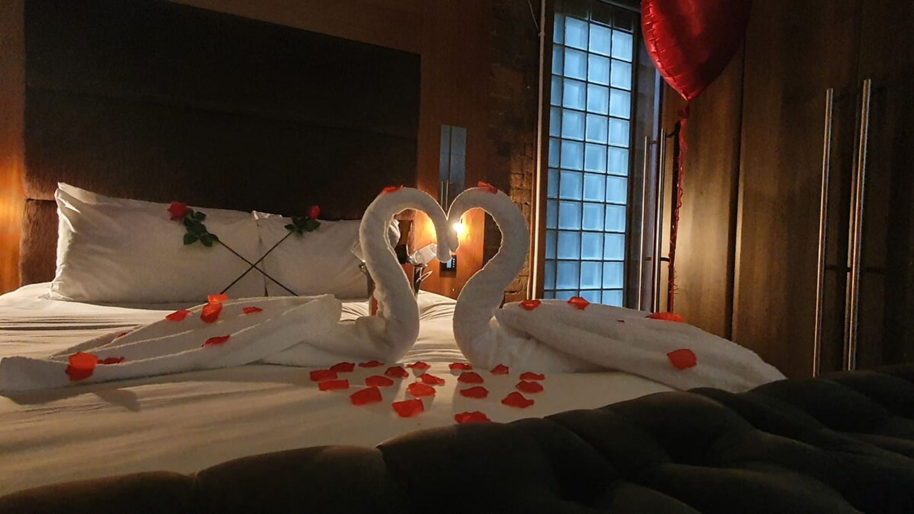 Una cama con sábanas blancas y pétalos de rosa, un arreglo de toallas con forma de corazón y un adorno con forma de corazón rojo.