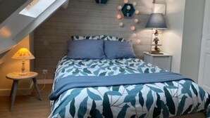 Une chambre avec un plafond en pente, un lit recouvert d’une couvre-lit à motifs de feuilles, une table de chevet avec une lampe et un luminaire fixé au mur.