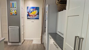 Une cuisine étroite avec des meubles blancs, un four encastré et un réfrigérateur. Deux tableaux sont accrochés au mur et un petit tapis recouvre le sol.