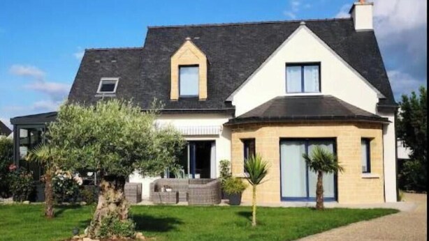 Une maison à deux étages, avec un toit sombre, une petite fenêtre de grenier et un porche à l’avant.