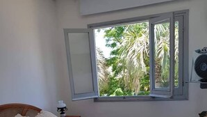 A bedroom with a bed, pillows, a nightstand, a window with a view of palm trees, and an air conditioning unit. - Hotel Puerto Las Palmas (Punta del Este)
