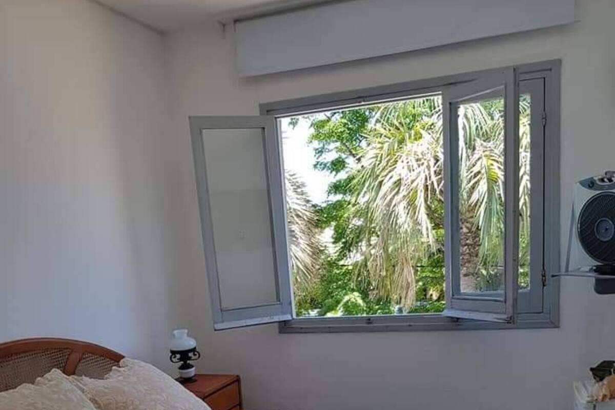 Quarto com cama, travesseiros, mesa de cabeceira, janela vista para palmeiras e ar condicionado.