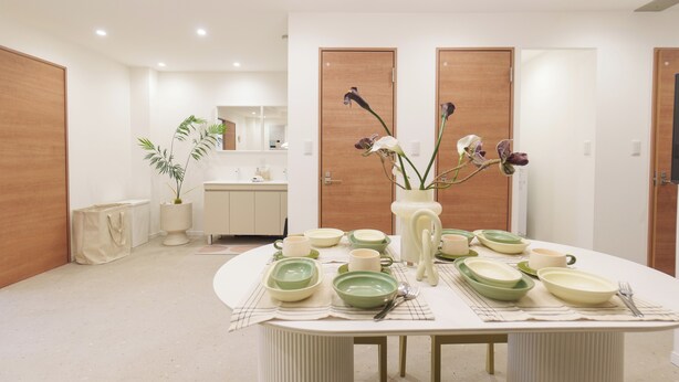 A modern dining area with a round table set for four, featuring green and white plates and bowls, a vase with flowers, and a placemat.