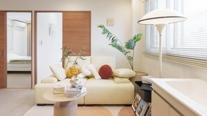 A modern living room with a beige sofa, a round coffee table, and a floor lamp. There is a window with blinds and a plant on the windowsill. - TORA HOTEL Sumidagawa (Tokyo)
