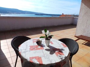 Ein Tisch mit Tischtuch, der Küchenutensilien, zwei Stühle und einen Blumentopf mit weißen Blüten auf einem Balkon mit Blick auf das Meer und die Berge zeigt.
