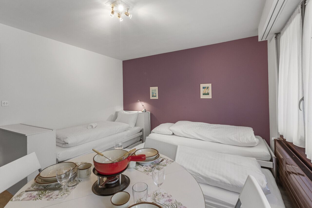 A small, clean room with two single beds, a table with a red pot and glasses, and a purple wall.